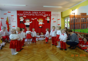 Na tle biało-czerwonej dekoracji stoją na przeciwko siebie ubrane na biało-czarwono dziewczynki i chłopcy. Dzieci przygotowują siędo tańca.